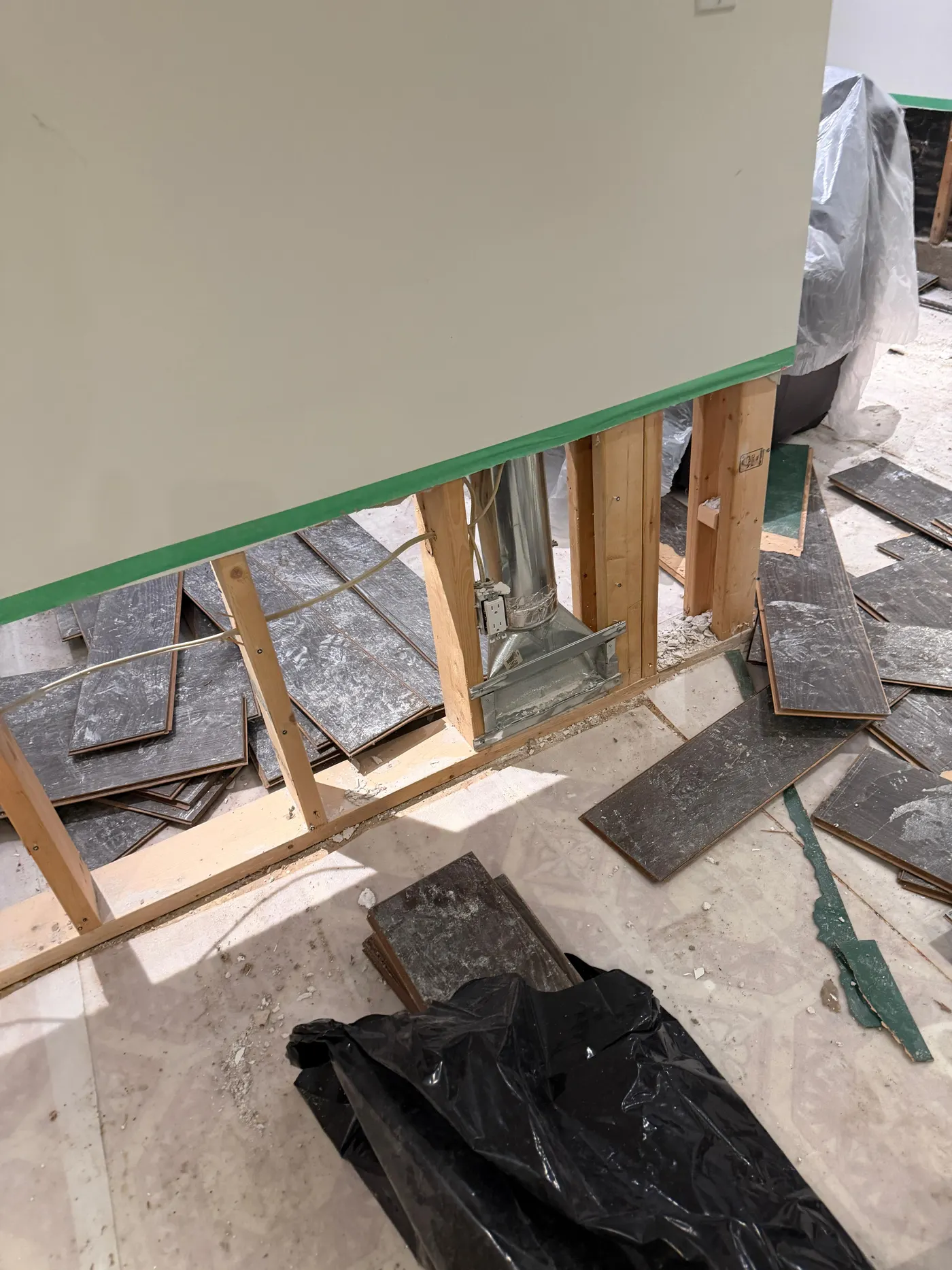 Lower section of a basement wall opened to the studs with laminate flooring lifted and stacked on the subfloor, ductwork visible inside the wall cavity